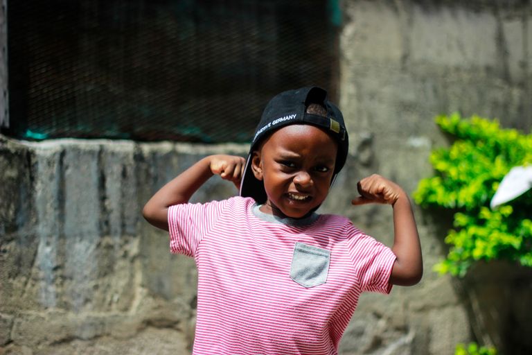 A young boy in a striped shirt poses confidently while flexing his muscles outdoors.