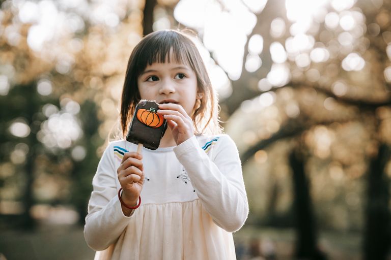 Adorable little girl in white clothes standing in park with bitten tasty chocolate Halloween sweet on stick and looking away