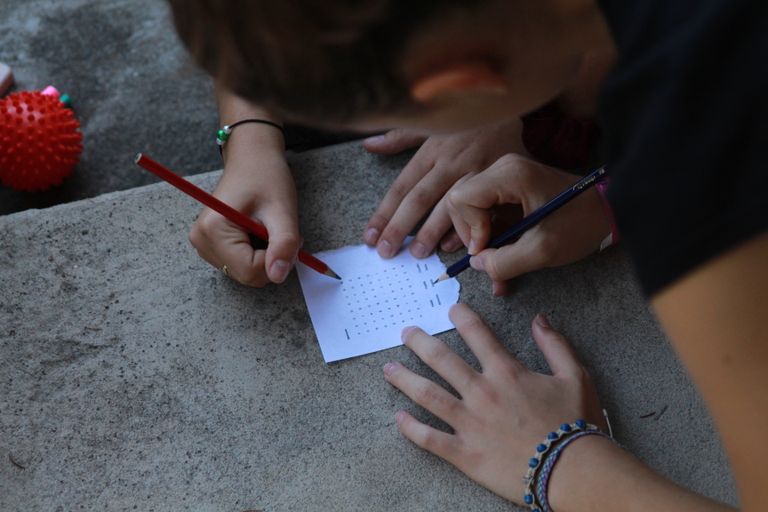 Close-up of hands solving a word puzzle outdoors using pencils on paper.
