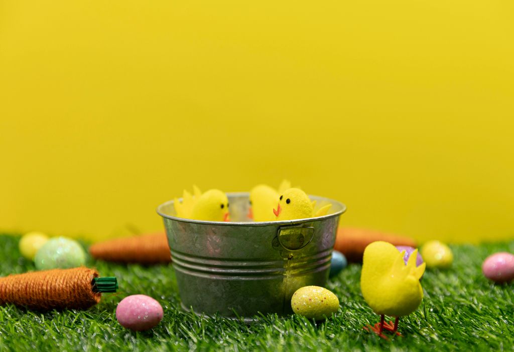 Vibrant Easter arrangement with toy chicks, eggs, and carrots on green grass against yellow background.